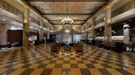 This magnificent hotel lobby is a masterclass in Beaux-Arts grandeur, offering a sweeping, voluminous space defined by soaring ceilings adorned with elaborately painted coffered panels featuring classical motifs in rich golds, reds, and blues. Suspended crystal chandeliers cast a warm, refined glow across the entire hall, complementing the ornate decorative plasterwork that frames the ceiling's perimeter.

Towering marble columns with veined golden and sienna tones line the space, supporting an upper-level mezzanine gallery framed by elegant ironwork balustrades. The signature checkerboard terrazzo floor in warm amber and deep brown tones anchors the space with timeless sophistication, stretching across every area of the lobby.

The central seating area features plush leather armchairs and curved sofas arranged around low tables, creating an inviting atmosphere for guests. Along the perimeter, intimate seating vignettes are appointed with console tables, decorative mirrors, and floral arrangements, adding warmth to the classical architecture. A sleek modern reception desk provides a subtle contemporary contrast.

Additional areas reveal an atmospheric lounge with dark-paneled walls, a water feature with river stones, and sculptural pendant installations. Throughout, the interplay of historic architectural detail and refined contemporary furnishings creates an atmosphere of understated luxury and timeless elegance.