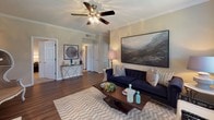 The Carlton-R floor plan offers a thoughtfully designed single-level layout with two bedrooms and two full bathrooms. The living room is a welcoming centerpiece, featuring hardwood-style flooring, crown molding, a ceiling fan with light fixture, and a fireplace, all bathed in natural light from large windows dressed with patterned curtains. The space flows seamlessly into a dedicated dining area, which is highlighted by a chandelier and a generous window overlooking greenery outside, creating a bright and inviting atmosphere for gatherings. The kitchen is well-appointed with ample light wood cabinetry, dark granite-style countertops, a double basin sink, and a full suite of appliances including a stainless steel range and dishwasher. Both bedrooms offer a calm, neutral palette with plush carpeting, large windows with outdoor views, and generous space for furnishings. Each bedroom is served by a full bathroom featuring a tub-shower combination with classic white tile accented by a bold dark tile border. Throughout the residence, warm tones, consistent finishes, and an open, connected layout combine to create a comfortable and cohesive living environment.