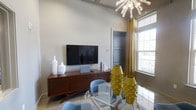 This thoughtfully designed single-floor space showcases a cohesive mid-century modern aesthetic that blends form and function seamlessly. The living area serves as a central focal point, anchored by a sleek wall-mounted flat-screen television atop a warm walnut-toned credenza with angled legs — a hallmark of retro-modern design. The space is elevated by a striking sputnik-style chandelier that draws the eye upward, hinting at the generous ceiling height and adding a sculptural, artistic quality to the room. Natural light pours in through large windows dressed with golden yellow curtains, creating a warm and inviting atmosphere. A glass-top dining table surrounded by powder-blue accent chairs introduces a playful yet refined contrast to the warm wood tones. Eye-catching golden textured vases serve as bold decorative statements, reinforcing the space's confident design sensibility. The neutral wall palette allows the curated furnishings and décor to take center stage, while the soft area rug underfoot adds texture and comfort. A dark-toned interior door with paneled detailing provides an elegant architectural accent. Overall, the space presents a well-balanced blend of retro inspiration and contemporary living.