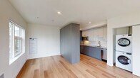 Located at 1341 N Killingsworth St in Portland's Piedmont neighborhood, this thoughtfully designed unit offers a clean, modern aesthetic throughout its single-floor layout. Light hardwood flooring flows seamlessly across the entire space, enhancing the sense of openness and continuity.

The open-concept living area is bright and airy, bathed in natural light from large windows that frame views of the surrounding neighborhood's mature trees and streetscape. A set of glass doors leads to a private balcony, further extending the sense of space.

The galley-style kitchen is efficiently designed with two-tone cabinetry — slate-blue lower cabinets paired with white upper cabinets — and stainless steel appliances including a refrigerator, microwave, and dishwasher. A full-size stacked washer and dryer unit is conveniently tucked nearby, adding practical functionality.

A full bathroom features light hardwood-style flooring, a combined soaking tub and shower, a sleek vanity with blue-gray cabinetry, an undermount sink, and a large mirror. The clean white walls and polished fixtures give the space a crisp, contemporary feel.

Recessed lighting throughout and a neutral white palette keep the interior feeling fresh and versatile, making this a well-appointed unit ideal for modern urban living in North Portland.