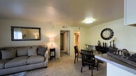 This single-floor unit showcases an open-concept layout that seamlessly integrates the living and dining areas into one cohesive, comfortable space. The living area features plush wall-to-wall carpeting in a neutral tone, complemented by warm beige walls that create an inviting atmosphere. Ceiling-mounted flush lighting provides consistent illumination throughout the space, while additional floor and table lamps contribute warm ambient light.

The dining area accommodates a round table with matching dark-stained wood chairs, positioned alongside a coordinating sideboard that offers additional storage and display space. A large decorative clock anchors the dining wall, adding a notable design element to the room.

From the main living space, two doorways are visible. One opens to a spacious walk-in closet featuring built-in shelving, offering generous storage capacity. The second provides access to a bathroom, which is partially visible and appears to include a vanity with mirror. The textured ceiling and consistent flooring throughout unify the space, contributing to an organized and functional single-level floor plan.