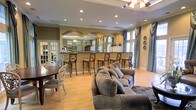 This single-floor space showcases a thoughtfully designed open-concept layout that seamlessly connects the living, dining, and kitchen areas. The interior is anchored by warm hardwood flooring that flows throughout, complemented by neutral olive-toned walls and crisp white crown molding that adds a refined architectural detail to the space.

A striking bar counter serves as a focal point, featuring a row of classic wooden barstools with upholstered seats. Behind the bar, an arched mirrored backsplash creates a sense of depth and elegance, reflecting light and visually expanding the space. Recessed lighting in the ceiling provides consistent, well-distributed illumination throughout the open area.

The living area is furnished with a generous, plush sofa, creating a comfortable gathering space, while a separate dining area with a round table and ornate white chairs adds a touch of classic styling. Large windows dressed with floor-length draping curtains in soft blue-gray tones allow ample natural light to filter into the space, enhancing its airy ambiance. Decorative wall accents and a statement chandelier visible in the mirrored reflection further elevate the overall aesthetic. The space strikes a balance between casual comfort and classic elegance throughout.