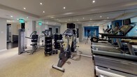 This fitness center is thoughtfully designed to support a comprehensive workout experience. The space is organized into distinct functional zones, beginning with a cardio area that lines one side of the room. Life Fitness elliptical trainers and a stationary cycle are positioned along floor-to-ceiling windows that overlook an indoor pool, providing an energizing visual connection to the aquatic facility beyond. Recessed lighting throughout the tray-ceilinged space ensures consistent, well-distributed illumination.

A mirrored wall extends along one section of the gym, visually expanding the area and providing functional feedback during strength training. A dedicated free weights zone features a full dumbbell rack alongside benches and cable equipment. Rubberized flooring throughout offers durability and comfort underfoot. A corridor leads deeper into the facility, accented by a bold teal feature wall that adds a dynamic visual element to the otherwise neutral, taupe-toned interior.

The entrance to the fitness center is accessible via a tiled corridor shared with pool access, marked by a glass door with "Fitness Center & Pool" signage. A small service counter with mosaic tile detailing greets visitors upon entry, reinforcing the polished, amenity-grade character of the space.