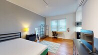This efficiently designed single-floor unit offers one bedroom, two bathrooms, and a kitchen, making smart use of its compact footprint. The bedroom serves as the largest room in the unit, featuring warm hardwood floors, neutral gray walls, and track lighting overhead. A large window with horizontal blinds fills the space with natural light, and a dedicated work desk area provides a functional workspace alongside open storage shelving. The kitchen is thoughtfully appointed with two-tone cabinetry — white upper cabinets paired with dark lower cabinets — complemented by light countertops, a stainless steel refrigerator, dishwasher, and built-in microwave. A small dining table with seating tucks neatly beside the kitchen, maximizing utility within a tight footprint. The bathroom features a full-height glass sliding shower enclosure with white subway tile, a toilet, and an open-shelf vanity over stone-look tile flooring, conveying a clean and modern aesthetic. A closet area provides additional storage with a hanging rod. Throughout the unit, hardwood flooring and crisp white trim create a cohesive, well-maintained look. The overall ambiance is practical, clean, and well-suited for individuals seeking a fully functional compact living space.