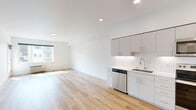 This thoughtfully designed one-bedroom, one-bathroom unit offers a clean, modern aesthetic throughout its well-utilized single-floor layout. The open-concept kitchen and living area serves as the heart of the unit, featuring light hardwood flooring that flows seamlessly across the space. The kitchen is outfitted with sleek white cabinetry extending to the ceiling for maximum storage, a white subway tile backsplash, light countertops, and a full suite of stainless steel appliances including a refrigerator, range, over-the-range microwave, and dishwasher. Recessed lighting illuminates the kitchen and living area, creating a bright, airy atmosphere complemented by natural light from a large window. The living area offers access to a private balcony through a glass-paned door, providing outdoor views of the surrounding neighborhood. The bedroom is a comfortable, well-proportioned room with its own window overlooking the balcony area and a ceiling fan for added comfort. The bathroom continues the modern, minimalist design with a large vanity featuring ample drawer and cabinet storage, a wide mirror with overhead lighting, and hardwood-style flooring. A stacked washer and dryer unit is conveniently located within the bathroom, adding practical in-unit laundry functionality.
