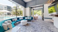 This outdoor amenity space is designed to deliver a resort-like experience within a residential community. At its center is a generously sized swimming pool with a distinctive island platform, surrounded by a wide paver deck lined with lounge chairs and shaded umbrellas. Tall pines, palms, and dense greenery frame the perimeter, creating a sense of seclusion and natural beauty.

The covered pavilion serves as the social hub of the space, featuring soaring ceilings painted in a cool slate blue and walls clad in white shiplap. A boldly curved teal sectional sofa anchors the lounge area around a sleek white round coffee table, offering a stylish and comfortable gathering spot. Large sliding glass panels open the pavilion entirely to the outdoors, blurring the line between interior and exterior. An orb chandelier adds an elegant finishing touch.

Adjacent to the lounge is an outdoor dining area with teak furniture, positioned next to a fully equipped grilling station with stainless steel grills — ideal for casual entertaining. A fire pit lounge with white Adirondack chairs provides an inviting spot for evening gatherings. Throughout, herringbone-patterned pavers unify the space with a refined, cohesive aesthetic that complements the modern multi-story residential buildings surrounding the courtyard.