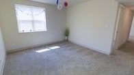 This single-storey space presents a clean, understated interior defined by its simplicity and functionality. The room showcased is finished with wall-to-wall carpet in a neutral beige-grey tone, providing a soft and comfortable underfoot feel throughout. The walls are painted in a light, warm off-white shade, contributing to an open and airy atmosphere.

A window fitted with horizontal blinds allows natural light to filter into the room, casting soft patches of sunlight across the carpeted floor and enhancing the sense of brightness. The window treatment provides both privacy and light control, making it a practical feature of the space.

The room benefits from a modest yet functional layout, with sufficient open floor space to accommodate a variety of furnishings. Decorative elements include colorful hanging paper ornaments suspended from above and a tall potted ornamental grass plant positioned in the corner, adding a subtle touch of character to the otherwise minimalist environment.

White baseboards run along the perimeter of the room, providing a clean finish where the walls meet the floor. Overall, the space offers a calm, neutral canvas well-suited for a range of uses.
