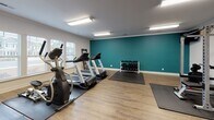 Located at 1601 Riddick Ln in Savannah, Georgia, this single-level fitness center offers a comprehensive workout environment designed to accommodate a wide range of exercise routines. The space is thoughtfully organized into distinct functional zones, allowing for both cardiovascular training and strength work without congestion.

The cardio area is positioned along a windowed wall, where multiple windows with white shutters fill the space with natural light. This section houses two treadmills, an elliptical trainer, and a stair climber, all placed on protective rubber mats atop light wood-look flooring that runs throughout the facility.

The strength training area features a full power rack with a bench and barbell setup, a multi-station cable machine, and a dedicated dumbbell rack with a full range of weights. Protective floor mats are positioned beneath all equipment. A wall-mounted mirror and a mounted television are also present in this zone, enhancing both functionality and motivation.

The interior is finished with clean white walls, with one accent wall painted in a bold teal hue, adding a vibrant focal point to the space. Crown molding and flush fluorescent lighting contribute to a polished, well-maintained atmosphere. French doors provide access to the facility, completing a practical and welcoming fitness environment.