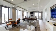 This single-floor space presents a well-appointed lounge and entertainment area with a refined contemporary design. The interior is defined by a coffered ceiling with recessed lighting, creating a structured yet open ambiance throughout. Floor-to-ceiling windows line multiple walls, flooding the space with natural light while offering sweeping panoramic city views, giving the room a distinctly elevated feel.

At the heart of the space, a sleek gas fireplace anchors a comfortable seating arrangement, with a wall-mounted television positioned above it for added functionality. White curved lounge chairs and a low round side table create an inviting conversation area, while a darker upholstered seating group provides an additional gathering zone nearby.

A shuffleboard game table serves as a central entertainment feature, reinforcing the social nature of the space. Toward one side, a dining area with a round table and dark wooden chairs offers a more intimate setting. The flooring transitions between large-format tile and area rugs, clearly delineating zones within the open layout. A glass door leads to what appears to be an outdoor terrace, further extending the space's connection to its urban surroundings.