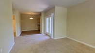 This single-floor residence offers a thoughtfully designed open layout that connects the living and kitchen areas in a seamless, functional flow. The living area is finished with plush wall-to-wall carpeting in a neutral tone, creating a warm and comfortable atmosphere throughout. Soft, light-colored walls complement the space, giving it a bright and airy feel.

A standout feature of the main living area is a set of elegant French doors, framed in crisp white trim, which introduce natural light and provide access to an adjacent area or outdoor space. The transition from the carpeted living area into the kitchen is marked by rich hardwood flooring, adding a distinct contrast and a touch of sophistication.

The kitchen is well-appointed with warm wood cabinetry and ample counter space, suggesting a practical and inviting cooking environment. A classic pendant light fixture hangs above the kitchen area, providing focused illumination and serving as a subtle decorative element. Clean white baseboards run throughout the space, tying together the overall aesthetic with a polished finish.

The open-plan design of this residence promotes a sense of spaciousness and ease of movement, making it well-suited for both everyday living and entertaining.