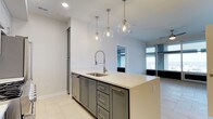 Located at 300 E Main St in the heart of Norfolk, Virginia, this single-floor residence offers a sleek, modern living experience defined by clean lines and high-end finishes throughout. The open-concept layout creates a seamless flow between the kitchen and living areas, maximizing the sense of space and functionality. The kitchen serves as a central highlight, anchored by a large island with a crisp white quartz countertop, a deep single-basin sink, and a professional-style spring-neck faucet. Slate-gray shaker cabinetry with brushed nickel hardware provides generous storage, while three globe pendant lights suspended from the ceiling add a contemporary, designer touch. The light-toned tile flooring extends throughout, enhancing the bright and airy ambiance. Large floor-to-ceiling windows in the adjacent living area invite abundant natural light and offer views of the surrounding cityscape, reinforcing the urban character of the space. Recessed lighting complements the natural light, ensuring a well-lit environment at any hour. The overall aesthetic is modern and refined, combining practical design elements with stylish details that reflect the energy of downtown Norfolk living.