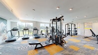 Located at 1400 Encore Place in Sanford, Florida, this fitness center offers a comprehensive and well-equipped workout environment designed to support a wide range of exercise routines. The single-floor facility is thoughtfully laid out, providing ample space for both cardio and strength training activities without feeling crowded.

The flooring features a striking patterned carpet tile design in grey and gold, adding a modern, energetic aesthetic throughout the space. Large floor-to-ceiling windows line the perimeter walls, flooding the interior with abundant natural light and offering open views of the surrounding greenery and landscape, creating an inviting atmosphere for workouts.

The cardio zone is well-stocked with multiple treadmills and elliptical trainers, all positioned to take advantage of the outdoor views. The strength training area includes a multi-station cable machine, a power rack with bench, adjustable benches, and a dedicated dumbbell rack, catering to a full range of resistance exercises. Wall-mounted flat-screen televisions are positioned for easy viewing during workouts.

Recessed ceiling lighting complements the natural light, ensuring the space remains bright and functional at all hours. The clean, neutral wall tones and open layout contribute to an uncluttered, professional environment that is both functional and welcoming for fitness enthusiasts of all levels.