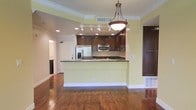 This single-floor space offers a thoughtfully designed open-concept layout that seamlessly connects the kitchen and living areas. The kitchen is a standout feature, showcasing rich dark wood cabinetry that extends to the upper walls, complemented by dark granite countertops that add a touch of elegance. Stainless steel appliances, including a French door refrigerator with an ice and water dispenser and a built-in microwave, provide a modern, functional touch. A raised breakfast bar peninsula defines the kitchen space while maintaining an open flow to the rest of the room. Overhead, a classic pendant light fixture anchors the kitchen area, while recessed lighting illuminates the space throughout. The warm, medium-toned hardwood flooring runs continuously across the open living area, adding richness and cohesion to the overall design. Soft yellow-green walls reflect light beautifully, contributing to a bright and inviting ambiance. Crown molding adds a refined architectural detail throughout. A hallway visible in the background hints at additional rooms beyond, rounding out this well-appointed and move-in-ready space.