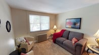 This single-floor residence offers a thoughtfully laid-out living space finished in warm, neutral tones that create a cohesive and inviting atmosphere throughout. The living room serves as the heart of the home, featuring soft carpeting, a classic cast-iron radiator, and a large window fitted with blinds that allows natural light to filter in while overlooking lush greenery outside. Wall-mounted artwork adds a pop of color to the understated palette.

The kitchen is efficiently designed with light maple cabinetry, dark laminate countertops, a stainless steel sink, and black appliances including an over-the-range microwave and a full-size refrigerator, providing all the essentials in a compact and functional layout.

The space includes two well-sized bedrooms, both carpeted and finished in the same warm neutral tones. Each room is equipped with a ceiling fan for added comfort, and multiple windows provide ample natural light and views of the surrounding greenery. One bedroom features a convenient open closet, while both rooms offer sufficient space for standard bedroom furnishings and storage.

The bathroom is clean and practical, featuring tile flooring, a pedestal sink, a bathtub with shower combination, and a window for natural ventilation. A cast-iron radiator provides consistent warmth. Overall, this residence offers a comfortable and functional living environment with consistent, classic finishes throughout.