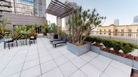 This expansive rooftop terrace offers a well-designed urban outdoor retreat with a variety of functional zones and thoughtful landscaping. The space is anchored by a striking overhead pergola that provides partial shade while framing views of the surrounding city skyline. Large-format porcelain tiles cover the main terrace floor, lending a clean, contemporary aesthetic throughout.

Generous galvanized metal planters are strategically positioned across the terrace, housing a diverse selection of tropical and ornamental plants, including tall yuccas, banana trees, bamboo, and colorful flowering ground cover. These lush plantings provide natural privacy screening and a verdant atmosphere that contrasts beautifully with the urban setting.

A dedicated lounge area features sleek reclining chaise lounges positioned beneath the pergola, ideal for relaxing and taking in the city views. Nearby, a circular built-in wooden bench surrounds a central table, shaded by a vibrant teal market umbrella and accented with bold black-and-white chevron cushions. A stainless steel gas grill is also available, making the terrace well-suited for outdoor dining and entertaining. The overall ambiance is modern, lively, and resort-inspired.