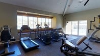 This fitness center offers a comprehensive workout environment designed to accommodate a variety of exercise routines. The space is thoughtfully laid out across a single floor, making it easy to navigate between different workout zones. Large multi-pane windows line one wall, flooding the room with natural light and providing views of the surrounding outdoor area, while a sliding glass door offers additional brightness and convenient access to the exterior. The ceiling features recessed lighting and subtle beam detailing, adding a clean, structured aesthetic to the room. The flooring is a combination of carpet and protective exercise mats, providing both comfort and functionality throughout the space. Cardio equipment is well-represented, with multiple treadmills and elliptical trainers positioned to take advantage of the natural light. A dedicated free weights area includes a fully stocked dumbbell rack with an organized range of weights, alongside a weight bench for strength training. Exercise balls and additional equipment round out the offerings. The overall ambiance is practical and motivating, with neutral wall tones and ample lighting creating an inviting atmosphere suited for both focused individual workouts and general fitness routines.