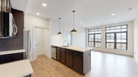 Located at 1050 N. 4th St in Columbus, Ohio's vibrant Short North neighborhood, this single-floor residence combines contemporary design with functional living. The open-concept kitchen serves as the heart of the space, showcasing a large central island with a white quartz countertop, undermount sink, and stainless steel dishwasher. Dark espresso cabinetry lines the walls, complemented by a classic white subway tile backsplash and a full suite of stainless steel appliances, including a refrigerator, range, and over-the-range microwave. Pendant lighting with black and gold fixtures adds a modern industrial accent overhead. In-unit laundry is conveniently tucked into the kitchen area. The living space flows naturally from the kitchen and is bathed in natural light from large, grid-paned black-framed windows that frame sweeping views of the Columbus cityscape. Light-toned hardwood-style flooring runs throughout, lending warmth and continuity to every room. The bedrooms are well-proportioned, with neutral walls, continued hardwood-style floors, and generous window placements offering urban views. The bathrooms are finished to a high standard, featuring large-format tile flooring, dark subway tile surrounds in the walk-in glass-enclosed showers, and dark-cabinetry vanities with white countertops.