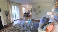 This single-floor residence offers an open, flowing layout that seamlessly connects the living and dining areas. The living room features rich hardwood flooring partially covered by a plush, textured area rug, anchoring a glass-topped coffee table with a sculptural chrome base. A white sofa with blue accent pillows provides comfortable seating, while decorative metallic wall plates add an artistic touch to the soft green walls. Large sliding glass doors flood the space with natural light and provide direct access to an outdoor area, complemented by elegant white patterned drapes. The open passage leads into a dedicated dining area, where a dark wood dining table and matching chairs sit beneath a classic pendant light fixture. A framed artwork on the wall adds a pop of color to the dining space, which also benefits from natural light through a window with blinds. The consistent hardwood flooring throughout both spaces creates visual continuity, while the neutral wall tones and carefully chosen accents contribute to a warm, welcoming ambiance suited for both everyday living and entertaining.