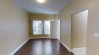 This single-floor space features a clean, functional design with warm neutral-toned walls that create an inviting atmosphere throughout. The main living area is highlighted by rich, dark hardwood flooring that adds a sense of warmth and continuity to the space. A window along the exterior wall allows natural light to filter in through vertical blinds, brightening the room and providing views of the surrounding outdoor area. The ceiling is smooth and well-proportioned, fitted with a flush-mount light fixture that provides even, ambient illumination. Clean white baseboards run along the perimeter of the room, offering a crisp contrast to the warm wall tones and dark flooring. A doorway with a classic six-panel white door connects to an adjacent room, suggesting a practical and well-organized floor plan. A recessed nook or entryway detail is visible along one wall, adding subtle architectural interest. The overall ambiance of the space is understated and neutral, making it highly adaptable for various furnishing styles and uses. The space presents a move-in ready condition with well-maintained finishes throughout.