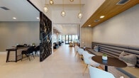 Located in Miramar, FL, this single-floor clubhouse at 4970 SW 124th Ave offers a sophisticated and contemporary community gathering space. The main lounge corridor features soaring ceilings, large-format light tile flooring, and elegant gold pendant globe lights that draw the eye through the space toward floor-to-ceiling glass doors opening to outdoor views of lush tropical landscaping. A striking laser-cut black decorative partition adds an artistic focal point at the entrance.

Seating areas are thoughtfully varied: a tufted gray banquette with round dark tables and modern white chairs provides casual dining-style seating, while plush blush armchairs with gold legs offer a relaxed lounge setting. A dedicated lounge nook showcases dramatic vertical wood slat wall and ceiling detailing paired with deep charcoal sectional sofas and globe pendant lighting.

The game room features exposed wood beam ceilings, a pool table, casual seating, and direct outdoor access, creating a vibrant social hub. A coffee and refreshment bar with warm wood cabinetry and open shelving serves the space conveniently. The leasing or administrative area includes a reception desk with a textured accent wall and mounted display. A private conference room is finished with wood-paneled walls, a large dining table, upholstered seating, and a bold sculptural brass chandelier.