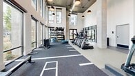 Located at 2490 Cobb Parkway SE in Atlanta, Georgia, this professionally equipped fitness center offers a generous, single-floor layout designed to accommodate a wide range of workout disciplines. The space is defined by its striking double-height ceilings and an abundance of natural light streaming through floor-to-ceiling and clerestory windows that line the perimeter, creating an open, energizing atmosphere.

The facility is organized into distinct functional zones. A dedicated cardio area features a row of treadmills, stair climbers, and other aerobic machines positioned alongside the expansive windows with views of the surrounding grounds. The free weights zone is well-appointed with a comprehensive dumbbell rack, weight benches, and cable machines, all set on durable rubberized flooring. A large open functional training area with a marked turf surface provides ample room for agility drills, sled pushes, and dynamic movement exercises. A separate glass-enclosed studio offers space for flexibility, stretching, and auxiliary training, with stability balls and mirrored walls.

Exposed concrete columns and industrial-style pendant lighting contribute to a modern aesthetic throughout. Large double doors connect the studio area to an outdoor green space, enhancing the facility's connection to the exterior environment. The overall design is clean, contemporary, and purpose-built for high-performance training.