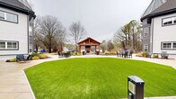 Located at 1551 Johnson Ferry Rd in Marietta, Georgia, this thoughtfully designed outdoor amenity space offers a welcoming and well-appointed environment for relaxation and social gathering. At the heart of the courtyard stands a handsome wood-framed pavilion with a gabled roof, housing an outdoor fireplace and mounted television, making it an ideal year-round gathering spot. The pavilion is surrounded by a broad stone patio furnished with multiple seating arrangements, providing ample space for residents or guests to gather and unwind. A meticulously maintained, vibrant green lawn anchors the center of the courtyard, framed by curved walkways and manicured landscaping beds filled with shrubs and seasonal plantings. A putting green is also present, adding a recreational touch to the grounds. The multi-story building adjacent to the courtyard features classic gray and white lap siding with dark trim accents, and ground-level sliding glass doors open directly onto the patio, seamlessly connecting indoor and outdoor spaces. Mature trees line the perimeter, providing a natural backdrop and a sense of privacy. The overall ambiance is polished yet approachable, offering a serene outdoor retreat in a residential Marietta community.