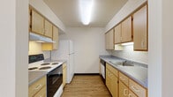 This single-floor unit offers a functional and straightforward layout with wood-look flooring extending throughout every room, creating a cohesive and easy-to-maintain interior. The galley-style kitchen runs along two parallel walls, outfitted with light wood cabinetry providing ample upper and lower storage, gray laminate countertops, a double-coil electric range, a full-size refrigerator, a dishwasher, and a stainless steel sink. Overhead fluorescent lighting keeps the kitchen well-illuminated.

Adjacent to the kitchen, the living area is open and bright, benefiting from a large window that invites natural light and offers views of surrounding greenery. A wall-mounted HVAC unit ensures climate control in this space. The entryway connects the main living area to the kitchen via an open passageway, with a front door featuring a secure lock and a built-in mailbox panel nearby.

The bedroom is a comfortable, well-proportioned room with a window providing natural light and an outdoor view, a baseboard heater, and an open closet with a hanging rod for storage. The bathroom includes a single-basin vanity with under-sink cabinetry, a lighted mirror, and a walk-in shower stall equipped with grab bars and a handheld showerhead.