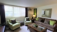 This single-floor space centers around a thoughtfully designed living room that balances comfort and style. The room features large windows dressed with vertical blinds and dark curtain panels, allowing for adjustable natural light while maintaining privacy. A wall-mounted air conditioning unit provides climate control convenience.

The seating arrangement includes a plush grey sofa adorned with decorative green and patterned throw pillows, complemented by two light green accent armchairs that introduce a refreshing pop of color. A woven ottoman adds a textural element alongside the chairs. The centrepiece of the seating area is a wood-framed glass coffee table, providing both visual openness and practical surface space.

Wall decor includes a framed green botanical artwork and a mosaic-style mirror, lending character to the neutral white walls. A warm table lamp on a dark side table contributes to a welcoming ambiance during evening hours. Carpeted flooring runs throughout, enhancing the room's comfort underfoot. Overall, the space presents a clean, inviting atmosphere with a cohesive green and grey color palette.