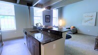 This two-floor model unit offers a thoughtfully designed open-concept layout that seamlessly blends functionality with contemporary style. The kitchen serves as a central focal point, featuring a large island with rich, dark granite countertops, an undermount sink, and dark wood cabinetry with brushed metal hardware — providing ample prep space and a natural gathering point. Pendant lighting above the island adds a warm, ambient glow that complements the overall aesthetic. Exposed ceiling beams lend an industrial character to the space, adding architectural interest and a sense of history to the interior. Large windows throughout the living area flood the space with generous natural light, creating a bright and welcoming atmosphere. The living area flows effortlessly from the kitchen, offering a comfortable and versatile arrangement suited for both relaxing and entertaining. Soft, neutral-toned carpeting anchors the living space, while the walls are finished in a cool, calming blue-gray palette. The two-floor configuration provides distinct zones for living and private use, enhancing the overall sense of space and livability throughout this well-appointed model unit.