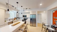 This single-floor residence is designed with a clean, contemporary aesthetic, characterized by light wood flooring, crisp white walls, and an open layout that promotes a seamless flow between living spaces. The kitchen serves as a central highlight, featuring flat-panel cabinetry in a light wood finish, subway tile backsplash, stainless steel appliances including a French door refrigerator, electric range, and built-in microwave. A central island with a white quartz countertop provides seating and additional workspace, while pendant lighting and track lighting illuminate the space effectively. The living area connects directly to a dedicated dining space, both benefiting from natural light and a cohesive, modern design. Two well-proportioned bedrooms offer natural light through large windows fitted with blinds and curtains, with views of lush landscaping including palm trees. The bathroom is finished with a wide vanity, dual gold-tone faucets, a white quartz countertop, and a glass-enclosed tiled shower. A hallway with decorative wall art leads to an in-unit stacked washer and dryer, adding everyday convenience to this well-appointed space.