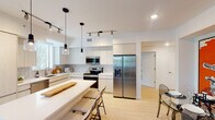 This single-floor residence is designed with a clean, contemporary aesthetic, characterized by light wood flooring, crisp white walls, and an open layout that promotes a seamless flow between living spaces. The kitchen serves as a central highlight, featuring flat-panel cabinetry in a light wood finish, subway tile backsplash, stainless steel appliances including a French door refrigerator, electric range, and built-in microwave. A central island with a white quartz countertop provides seating and additional workspace, while pendant lighting and track lighting illuminate the space effectively. The living area connects directly to a dedicated dining space, both benefiting from natural light and a cohesive, modern design. Two well-proportioned bedrooms offer natural light through large windows fitted with blinds and curtains, with views of lush landscaping including palm trees. The bathroom is finished with a wide vanity, dual gold-tone faucets, a white quartz countertop, and a glass-enclosed tiled shower. A hallway with decorative wall art leads to an in-unit stacked washer and dryer, adding everyday convenience to this well-appointed space.