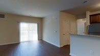 This single-floor residence offers an open and functional layout centered around a bright living area. The space features dark hardwood-style flooring that runs continuously throughout, providing a warm contrast against the neutral beige walls and crisp white baseboards and door trim. A large sliding glass door fitted with vertical blinds serves as the primary source of natural light, illuminating the living area and offering a view of the outdoor surroundings.

The open layout connects the living area to adjacent spaces, with a partial view of a kitchen counter featuring stone-look countertops visible to one side. Two paneled interior doors are positioned at the far end of the room, likely leading to bedrooms or utility spaces, and are framed with clean white trim that complements the overall neutral color palette. Ceiling height is standard, contributing to a comfortable and well-proportioned feel throughout. Recessed ceiling vents indicate a central HVAC system for year-round climate control. The overall ambiance is clean, minimal, and move-in ready, well-suited for a variety of furnishing styles.