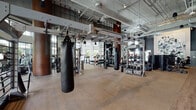 Located at 3200 Paseo Village Way in San Diego, CA, this state-of-the-art fitness center delivers a comprehensive workout experience within a thoughtfully designed, single-level facility. The space showcases an industrial-modern aesthetic, highlighted by exposed ductwork, soaring ceilings, and an abundance of natural light streaming through large, steel-framed windows that frame views of the surrounding outdoor greenery and palm trees.

The main workout floor is expansive and open, featuring rubber flooring and a wide array of strength and conditioning equipment, including heavy punching bags, cable machines, medicine ball racks, kettle bells, and a full dumbbell rack. A dedicated functional training area provides ample room for dynamic movement and group exercises. Cardio equipment, including a row of treadmills and rowing machines, is positioned along the perimeter near floor-to-ceiling windows, offering energizing outdoor views during workouts.

A connected lounge area transitions seamlessly between indoor and outdoor spaces, featuring a turf zone alongside a casual seating nook with artwork and a front desk or service counter. Bold graphic murals and artistic wall treatments add visual character throughout, while full-length mirrors and industrial pendant lighting reinforce the modern, motivating atmosphere. The facility is designed to accommodate a wide range of fitness activities in a polished, well-equipped environment.