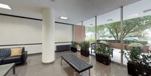 This single-floor commercial lobby presents a professional and welcoming environment defined by clean architectural lines and thoughtful design details. The main entrance features glass doors with transom windows, flooding the space with natural light and offering views of mature trees and the surrounding exterior. A polished terrazzo floor with geometric triangle inlay patterns anchors the entry and reception area, adding a refined decorative touch underfoot.

A dark wood reception desk serves as the central focal point upon entry, flanked by cylindrical column accents and accented with potted greenery that adds warmth to the neutral color palette. Recessed and flush-panel ceiling lighting ensures the space is evenly and brightly illuminated throughout.

A dedicated lounge area offers comfortable black leather seating — including a sofa, accent chairs, and a tufted bench — arranged around a glass-top coffee table. Framed artwork and tall indoor plants contribute to the space's polished aesthetic.

Toward the rear, a raised landing with low staircase steps leads to a bank of individual mailboxes, providing a practical utility zone integrated seamlessly into the overall design. Decorative vases and architectural column detailing echo the consistent style found throughout, making this lobby a functional yet visually cohesive space.
