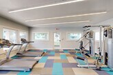 Located at 8135 N 34th Ave in Phoenix, Arizona, this fitness center offers a dedicated exercise space designed to support a well-rounded workout routine. The single-room facility is thoughtfully equipped with multiple True treadmills, an elliptical machine, and a multi-station strength training cable machine, providing options for both cardiovascular and resistance training.

The interior design strikes a clean, modern aesthetic with light gray walls and a distinctive geometric carpet in a mix of teal, blue, gray, and tan tones that adds visual energy to the space. Overhead, linear LED lighting runs across the ceiling, ensuring bright, even illumination throughout. A wall-mounted mini-split HVAC unit keeps the environment comfortable year-round.

Multiple white-framed windows line the walls, allowing natural light to flow in and offering views of the outdoor pool and surrounding property. A glass-paneled entry door features the facility's logo, contributing to a polished, branded appearance. An additional connected hallway suggests access to supplementary amenities nearby.

The layout is open and uncluttered, allowing users to move freely between equipment. Overall, this fitness center delivers a functional, modern, and inviting environment well-suited for daily exercise in the heart of Phoenix.