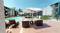 Located at 9931 Lurline Ave in the Chatsworth neighborhood of Los Angeles, this outdoor recreational space serves as a central amenity within a residential community. The area is defined by a generously sized swimming pool with clear blue water, complemented by an adjacent spa, both finished with classic tile surrounds and equipped with easy-access handrails. The pool deck is finished in a durable, textured aggregate surface that extends across the entire area, providing ample space for lounging and socializing.

Scattered throughout the deck are well-appointed seating arrangements, including wicker lounge chairs and deep-seated armchairs grouped beneath large canvas market umbrellas, offering shaded retreats from the Southern California sun. A row of chaise lounges lines one side of the pool area, ideal for sunbathing. Raised planter beds filled with tropical foliage add greenery and visual structure to the space.

The surrounding architecture features stucco-clad residential buildings in teal and white tones, with balconied units overlooking the courtyard. A single-story clubhouse or amenity building with a terracotta tile roof and floor-to-ceiling glass entry doors anchors one end of the pool area. Tall palm trees frame the perimeter, reinforcing the resort-like ambiance under the clear Los Angeles sky.