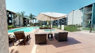 Located at 9931 Lurline Ave in the Chatsworth neighborhood of Los Angeles, this outdoor recreational space serves as a central amenity within a residential community. The area is defined by a generously sized swimming pool with clear blue water, complemented by an adjacent spa, both finished with classic tile surrounds and equipped with easy-access handrails. The pool deck is finished in a durable, textured aggregate surface that extends across the entire area, providing ample space for lounging and socializing.

Scattered throughout the deck are well-appointed seating arrangements, including wicker lounge chairs and deep-seated armchairs grouped beneath large canvas market umbrellas, offering shaded retreats from the Southern California sun. A row of chaise lounges lines one side of the pool area, ideal for sunbathing. Raised planter beds filled with tropical foliage add greenery and visual structure to the space.

The surrounding architecture features stucco-clad residential buildings in teal and white tones, with balconied units overlooking the courtyard. A single-story clubhouse or amenity building with a terracotta tile roof and floor-to-ceiling glass entry doors anchors one end of the pool area. Tall palm trees frame the perimeter, reinforcing the resort-like ambiance under the clear Los Angeles sky.