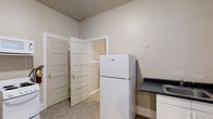 This single-floor space offers a straightforward and functional layout designed to make efficient use of every area. The kitchen is equipped with a full-size top-freezer refrigerator, dark granite-style countertops, and a sink, all set against light-toned walls and herringbone-patterned flooring. Classic five-panel white doors lead to additional areas of the unit, adding a traditional architectural touch. The living and sleeping areas feature wall-to-wall carpeting in a neutral gray tone, providing a comfortable underfoot feel. Large windows fitted with window-unit air conditioners allow for natural light while maintaining climate control throughout the space. Wide white baseboards run along the perimeter of each room, complementing the neutral beige and gray wall tones. The overall ambiance is clean and understated, offering a blank canvas ready for personalization. The open doorways between rooms create a connected flow, making the space feel cohesive. This unit is well-suited for those seeking a practical, move-in-ready space with functional essentials already in place.