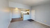 This single-floor residence offers an open-concept layout that seamlessly connects the living, dining, and kitchen areas. The living space is finished with plush grey carpeting, while the adjacent dining area transitions to smooth hardwood-style flooring, adding visual contrast and definition between the two zones.

The kitchen is centrally positioned and features a peninsula with a raised bar-height counter, providing both a functional workspace and a natural divider between the cooking and living areas. White cabinetry offers ample storage, complemented by stainless steel appliances including a refrigerator and microwave. Under-cabinet task lighting ensures a well-lit cooking environment.

The dining area is accented by a decorative chandelier, adding a touch of elegance to the otherwise clean and understated aesthetic. Soft grey walls run throughout the space, contributing to a calm, neutral ambiance that allows for versatile interior styling. White trim and baseboards provide crisp contrast against the walls. The layout is efficient and practical, making excellent use of the available floor space across the single level.
