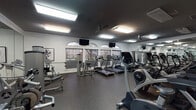 Located at 3535 E 10th St in Greenville, NC, this single-floor amenity center offers a thoughtfully designed collection of spaces for fitness, work, and social gathering. The expansive fitness area is equipped with Cybex cardio machines including ellipticals, treadmills, and stationary bikes, complemented by strength training equipment. Floor-to-ceiling mirrors, rubber flooring, ceiling fans, and arched windows that bring in natural light and outdoor views complete the workout environment. Adjacent to the gym, a dedicated lounge area features curved, contemporary sofas set against a bold typographic accent wall, exposed brick, and a striking multi-bulb chandelier, creating a dynamic social atmosphere. A cozy seating nook with leather chairs, teal textured wallcovering, and curated wall art provides an intimate gathering space. The dining area offers a wood table surrounded by vibrant orange chairs beneath industrial-style pendant lighting, with patterned wallcovering as a backdrop. The business center includes multiple computer workstations set against a bold teal patterned wall, with vibrant yellow-green carpet and flexible seating throughout. A café-style banquette area with marble-top tables and globe pendant lights rounds out this well-rounded and visually engaging community facility.