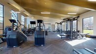 This well-appointed fitness center is designed to support a full range of workout routines across multiple distinct zones. The main training floor features soaring vaulted ceilings with warm wood paneling and exposed beam detailing, lending the space an open, airy quality. Herringbone-patterned carpet tiles cover the floor throughout the primary areas, while the dedicated studio room features rich hardwood flooring. Large, floor-to-ceiling windows line the walls, flooding the interior with natural light and offering views of the surrounding outdoor greenery.

The main floor is comprehensively equipped with a robust selection of strength training machines, cable systems, free weights, and dumbbell racks organized along mirrored walls. A row of treadmills and cardio machines is positioned to take advantage of the natural light and outdoor views. Wall-mounted televisions are distributed throughout the space for entertainment during workouts.

A separate studio room, accessible through glass-panel garage-style doors that open to the outdoors, houses rowing machines and spin bikes alongside a wall-mounted display screen. A water fountain is conveniently located near the transition between zones. The facility's thoughtful layout, generous scale, and high-end equipment make it a comprehensive and inviting fitness destination.