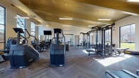 This well-appointed fitness center is designed to support a full range of workout routines across multiple distinct zones. The main training floor features soaring vaulted ceilings with warm wood paneling and exposed beam detailing, lending the space an open, airy quality. Herringbone-patterned carpet tiles cover the floor throughout the primary areas, while the dedicated studio room features rich hardwood flooring. Large, floor-to-ceiling windows line the walls, flooding the interior with natural light and offering views of the surrounding outdoor greenery.

The main floor is comprehensively equipped with a robust selection of strength training machines, cable systems, free weights, and dumbbell racks organized along mirrored walls. A row of treadmills and cardio machines is positioned to take advantage of the natural light and outdoor views. Wall-mounted televisions are distributed throughout the space for entertainment during workouts.

A separate studio room, accessible through glass-panel garage-style doors that open to the outdoors, houses rowing machines and spin bikes alongside a wall-mounted display screen. A water fountain is conveniently located near the transition between zones. The facility's thoughtful layout, generous scale, and high-end equipment make it a comprehensive and inviting fitness destination.