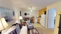 This single-floor unit offers a thoughtfully designed layout that maximizes its intimate scale. The open-concept living and dining area serves as the heart of the space, featuring neutral carpet flooring, soft neutral walls, and a ceiling fan with integrated lighting. A plush sofa, floral accent chair, and a small bistro-style dining table for two create a welcoming, functional arrangement. Natural light filters in through a sliding glass door dressed with sheer curtains, adding brightness to the living area.

The kitchen sits adjacent to the living space and is equipped with warm honey-toned wood cabinetry providing ample storage, a coil-burner electric range, a dishwasher, and a full-size refrigerator. The tile flooring transitions seamlessly from the kitchen into the dining area.

The bedroom offers a relaxed retreat, featuring carpeted flooring, a ceiling fan, and a window with dark curtain panels that allow for light control. Nautical-themed shadow box wall art adds a charming coastal touch to the space.

The full bathroom includes a tub-shower combination with white tile surround, a single-sink vanity with wood cabinetry, a large mirror, and a well-lit vanity light bar. A closet accessible from the main living area provides additional storage throughout the unit.