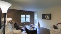 This single-floor space offers a welcoming and well-appointed living area designed with both comfort and style in mind. The living room serves as the focal point of the space, showcasing rich, dark hardwood flooring that runs throughout and provides a warm, grounded aesthetic. The walls are finished in a deep charcoal-brown accent tone, complemented by crisp white trim and baseboards that create a clean, contrasting look.

Natural light filters in through a double-pane window dressed with dark valance curtains, offering a view of lush greenery outdoors. A white French door with glass panel inserts adds architectural interest while allowing additional light into the room and providing access to the exterior. A wall-mounted flat-screen television is positioned prominently on the lighter accent wall, making it a functional centerpiece for the living area.

The room is furnished with a light-colored sofa accented with dark throw pillows, paired with a dark wood coffee table that anchors the seating arrangement. Tall decorative floor vases near the French door add a subtle artistic touch. The smooth, light-colored ceiling enhances the sense of openness throughout the space. Overall, the space presents a balanced blend of dark and light tones, creating an inviting and modern atmosphere.