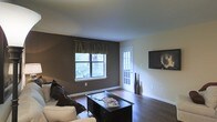 This single-floor space offers a welcoming and well-appointed living area designed with both comfort and style in mind. The living room serves as the focal point of the space, showcasing rich, dark hardwood flooring that runs throughout and provides a warm, grounded aesthetic. The walls are finished in a deep charcoal-brown accent tone, complemented by crisp white trim and baseboards that create a clean, contrasting look.

Natural light filters in through a double-pane window dressed with dark valance curtains, offering a view of lush greenery outdoors. A white French door with glass panel inserts adds architectural interest while allowing additional light into the room and providing access to the exterior. A wall-mounted flat-screen television is positioned prominently on the lighter accent wall, making it a functional centerpiece for the living area.

The room is furnished with a light-colored sofa accented with dark throw pillows, paired with a dark wood coffee table that anchors the seating arrangement. Tall decorative floor vases near the French door add a subtle artistic touch. The smooth, light-colored ceiling enhances the sense of openness throughout the space. Overall, the space presents a balanced blend of dark and light tones, creating an inviting and modern atmosphere.