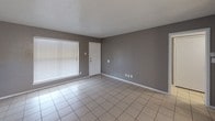 This single-story unit features a consistent, practical design throughout, with light-toned ceramic tile flooring laid in a grid pattern across every room. The walls are painted in neutral gray-taupe tones, complemented by white baseboards and textured popcorn ceilings that are consistent throughout the space.

The living area is generously sized, receiving natural light through large windows fitted with horizontal blinds. A front entry door opens directly into this open space, which flows toward the rest of the unit. An adjacent area, suitable for dining, connects to the kitchen.

The kitchen is designed with functionality in mind, featuring a peninsula with a granite-look countertop that provides seating and serves as a visual divider from the living and dining areas. White cabinetry lines the upper and lower walls, and the kitchen is equipped with a black refrigerator and black gas range. A window above the sink allows additional natural light into the workspace.

The bedrooms are well-proportioned, each featuring the same tile flooring, neutral walls, a window with blinds, and flush-mount ceiling lighting. The bathroom includes a white tub and shower combination, a toilet, and a vanity with a granite-look countertop, all finished with clean, simple fixtures in brushed chrome.