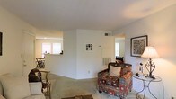 This single-story space presents an open, flowing layout that connects the living area to adjoining rooms, creating a sense of continuity throughout. The living room serves as the central hub, featuring neutral white walls and light carpeted flooring that contribute to a clean, understated aesthetic. Seating includes a patterned armchair with red, brown, and beige tones, complemented by additional wooden accent chairs, lending the space a casual yet functional character.

A central partial wall divides the space without fully enclosing it, maintaining an open feel while subtly defining separate zones. A built-in HVAC unit is integrated into this dividing wall, providing climate control. Ceiling vents are also visible, indicating a supplementary ventilation system throughout.

Natural light filters in through multiple windows, including those visible in what appears to be a dining area to the left, where a small table and chairs can be seen. An additional window to the right brightens a secondary area, possibly a bedroom or hallway. A framed artwork piece adorns the right wall, adding a modest decorative touch. Overall, the space offers a practical, livable environment with a neutral palette and connected, multi-functional areas.