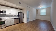 This single-floor unit is thoughtfully designed with a functional open-concept layout that integrates the kitchen and living areas seamlessly. The kitchen is well-appointed with a full suite of stainless steel appliances, including a refrigerator, range with oven, built-in microwave, and dishwasher. White shaker-style cabinetry extends to the upper and lower walls, complemented by a neutral countertop and gray tile backsplash. Track lighting overhead provides focused illumination throughout the cooking space. The kitchen flows directly into the living area, which features durable hardwood-style flooring and a decorative textured ceiling that adds visual character. Wainscoting panels line the lower portion of the living room walls, lending a classic architectural detail. A window allows natural light to brighten the space. A separate carpeted bedroom offers a quieter retreat, also featuring wainscoting and a window with blinds. The bathroom is finished in a clean, contemporary style with large-format gray tile flooring, a white vanity with black hardware, a tiled tub-shower combination, and recessed lighting, creating a bright and polished feel throughout.