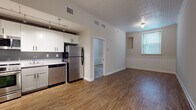 This single-floor unit is thoughtfully designed with a functional open-concept layout that integrates the kitchen and living areas seamlessly. The kitchen is well-appointed with a full suite of stainless steel appliances, including a refrigerator, range with oven, built-in microwave, and dishwasher. White shaker-style cabinetry extends to the upper and lower walls, complemented by a neutral countertop and gray tile backsplash. Track lighting overhead provides focused illumination throughout the cooking space. The kitchen flows directly into the living area, which features durable hardwood-style flooring and a decorative textured ceiling that adds visual character. Wainscoting panels line the lower portion of the living room walls, lending a classic architectural detail. A window allows natural light to brighten the space. A separate carpeted bedroom offers a quieter retreat, also featuring wainscoting and a window with blinds. The bathroom is finished in a clean, contemporary style with large-format gray tile flooring, a white vanity with black hardware, a tiled tub-shower combination, and recessed lighting, creating a bright and polished feel throughout.