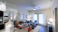 This single-floor space features an open-concept layout that seamlessly connects the dining and living areas, creating a cohesive and functional environment. The living area is anchored by a comfortable gray sofa accented with blue and neutral throw pillows, complemented by a dark-finish coffee table atop a plush white area rug. Dark hardwood-style flooring runs throughout, providing a clean contrast to the light-toned walls. A ceiling fan with an integrated light fixture is centrally positioned overhead, enhancing both airflow and ambient lighting. Large windows dressed with sheer white curtains and horizontal blinds allow abundant natural light to fill the room, while a glass-paneled door provides access to an outdoor area. The dining space includes a round white table paired with distinctively styled orange chairs, adding a pop of color and personality. Framed artwork on the walls contributes to the polished, contemporary aesthetic. A large mirror near the dining area visually expands the space, reinforcing the open and airy atmosphere throughout.
